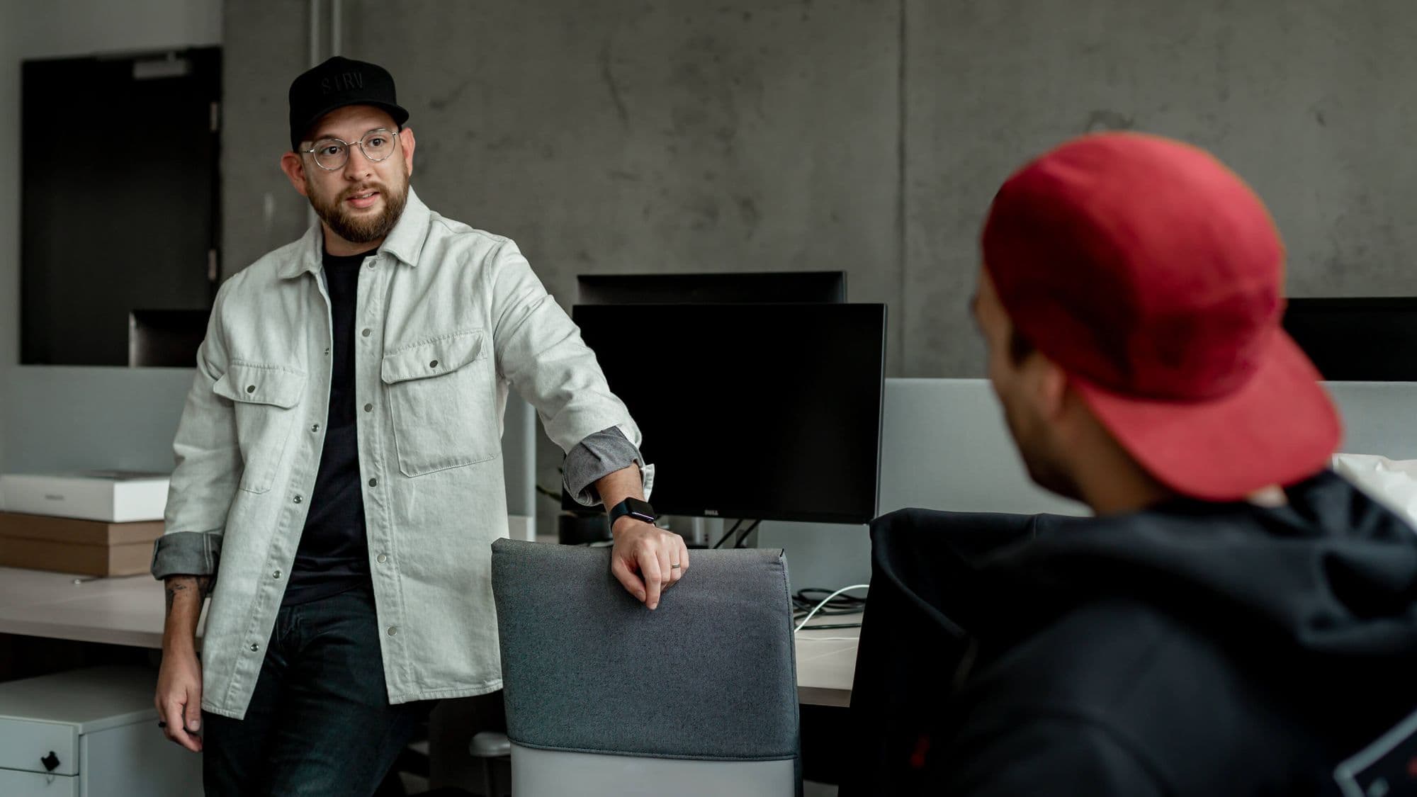 A man in a light jacket leans on a chair, speaking to a person wearing a red cap, in a modern office setting.