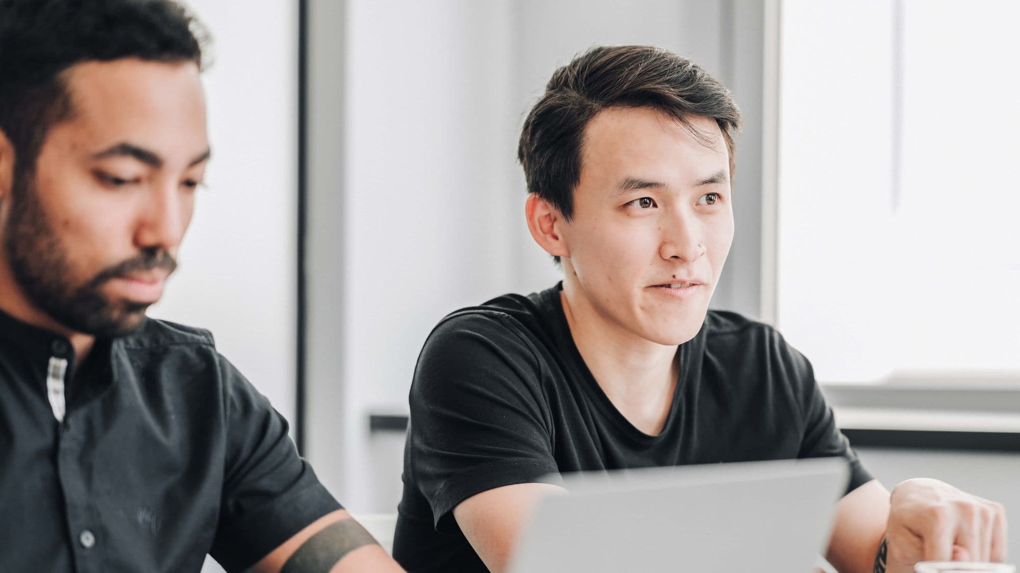 Two men engaged in a discussion at a table, with one looking intently at the other, who is typing on a laptop.