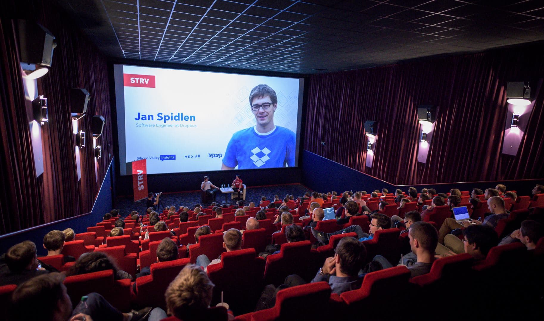 A speaker presents on stage in a theater, with an audience engaged and a large screen displaying Jan Spidlen from Dropbox.