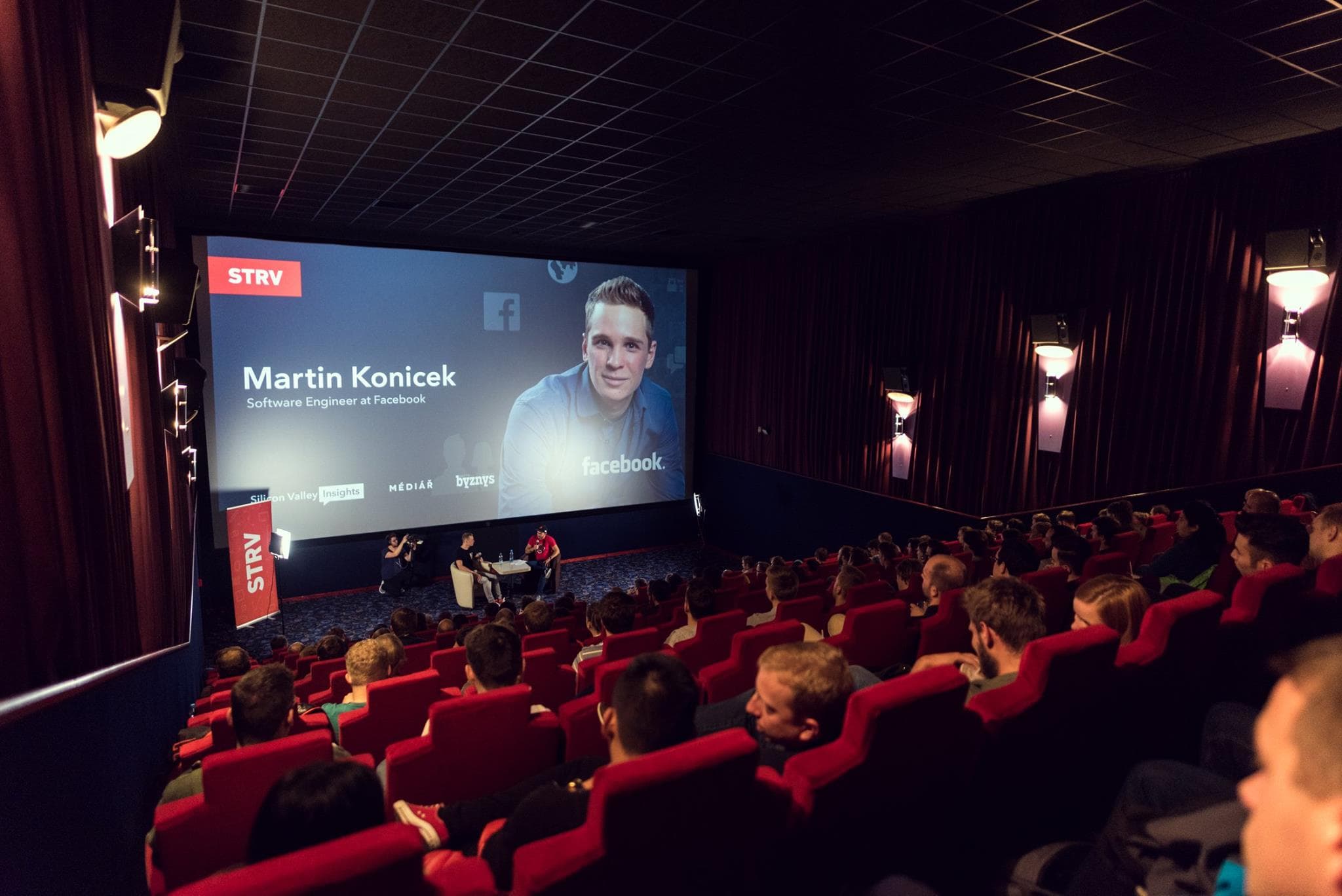 A crowded theater with red seats features Martin Konicek, a Facebook software engineer, speaking at an event.