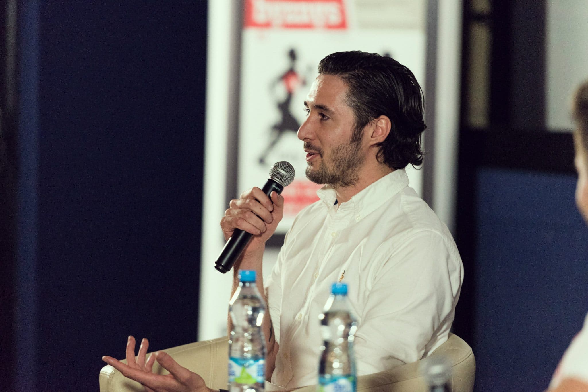 A man in a white shirt speaks into a microphone during a discussion, seated with water bottles on a table.