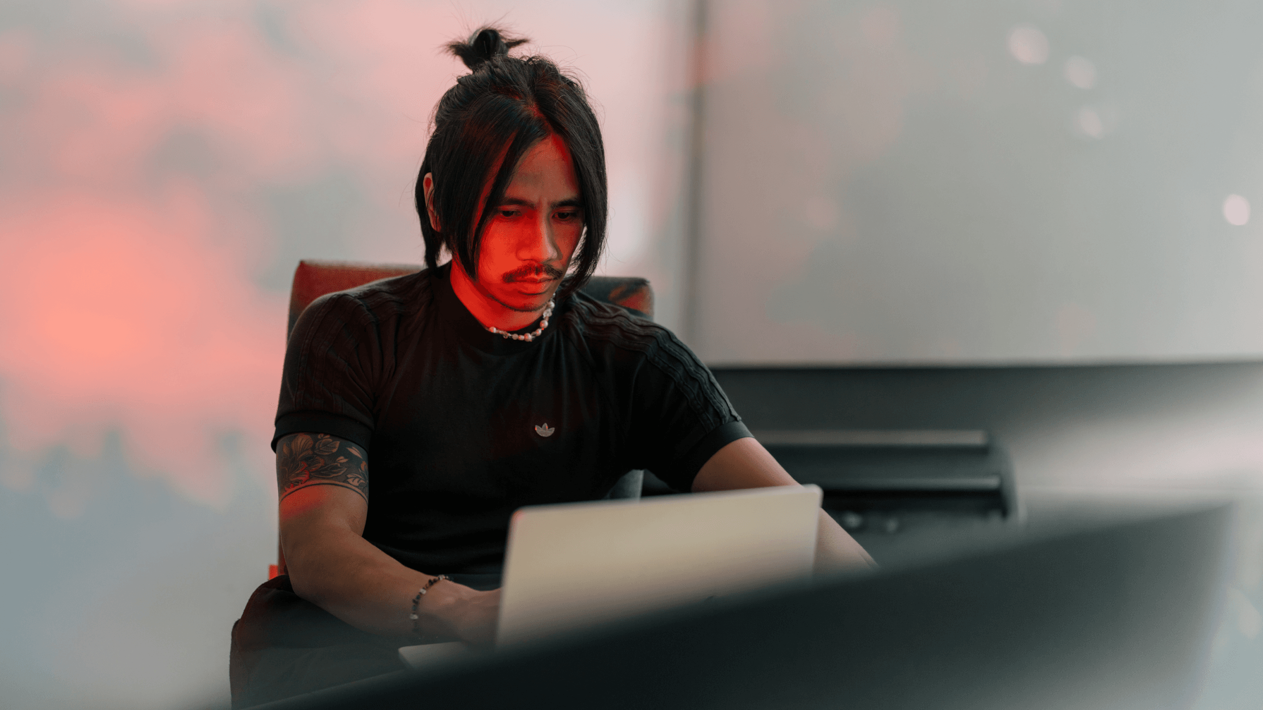 A person with long hair styled in a bun is focused on a laptop, seated in a dimly lit room with red lighting.